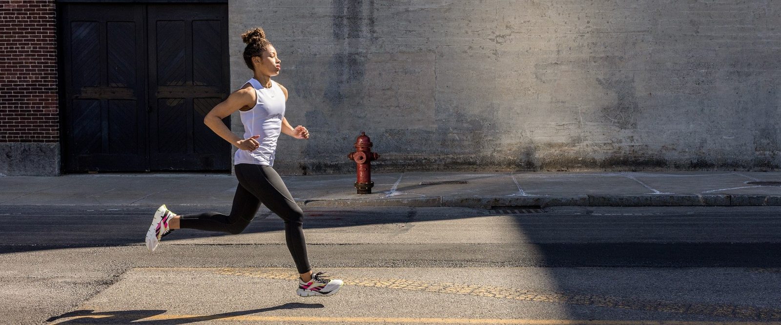 FCSI | A person in athletic wear is running on an urban street. They are wearing a white tank top, black leggings, and running shoes. The background features a brick wall, a gray concrete wall, a red fire hydrant, and shadows cast on the street.