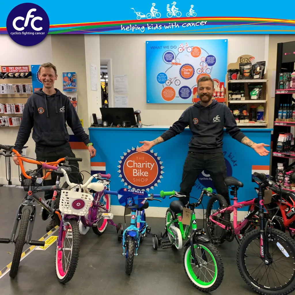 FCSI | Two people stand behind a row of colorful children's bikes inside a bike shop. A blue counter with a "The Charity Bike Shop" sign is in the background, along with a wall display. CFC logo is at the top, and shelves of bike accessories are visible.