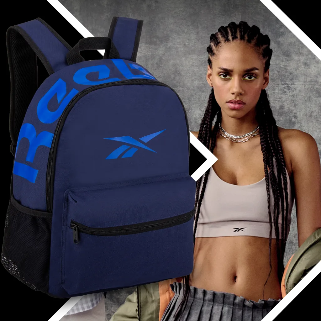 FCSI | A young woman with braided hair wears a beige sports bra and layered necklaces. She stands against a textured gray background. In the foreground, there is a navy blue Reebok backpack with bold blue lettering.