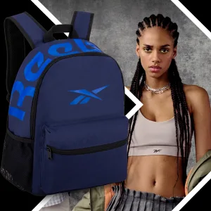 FCSI | A young woman with braided hair wears a beige sports bra and layered necklaces. She stands against a textured gray background. In the foreground, there is a navy blue Reebok backpack with bold blue lettering.
