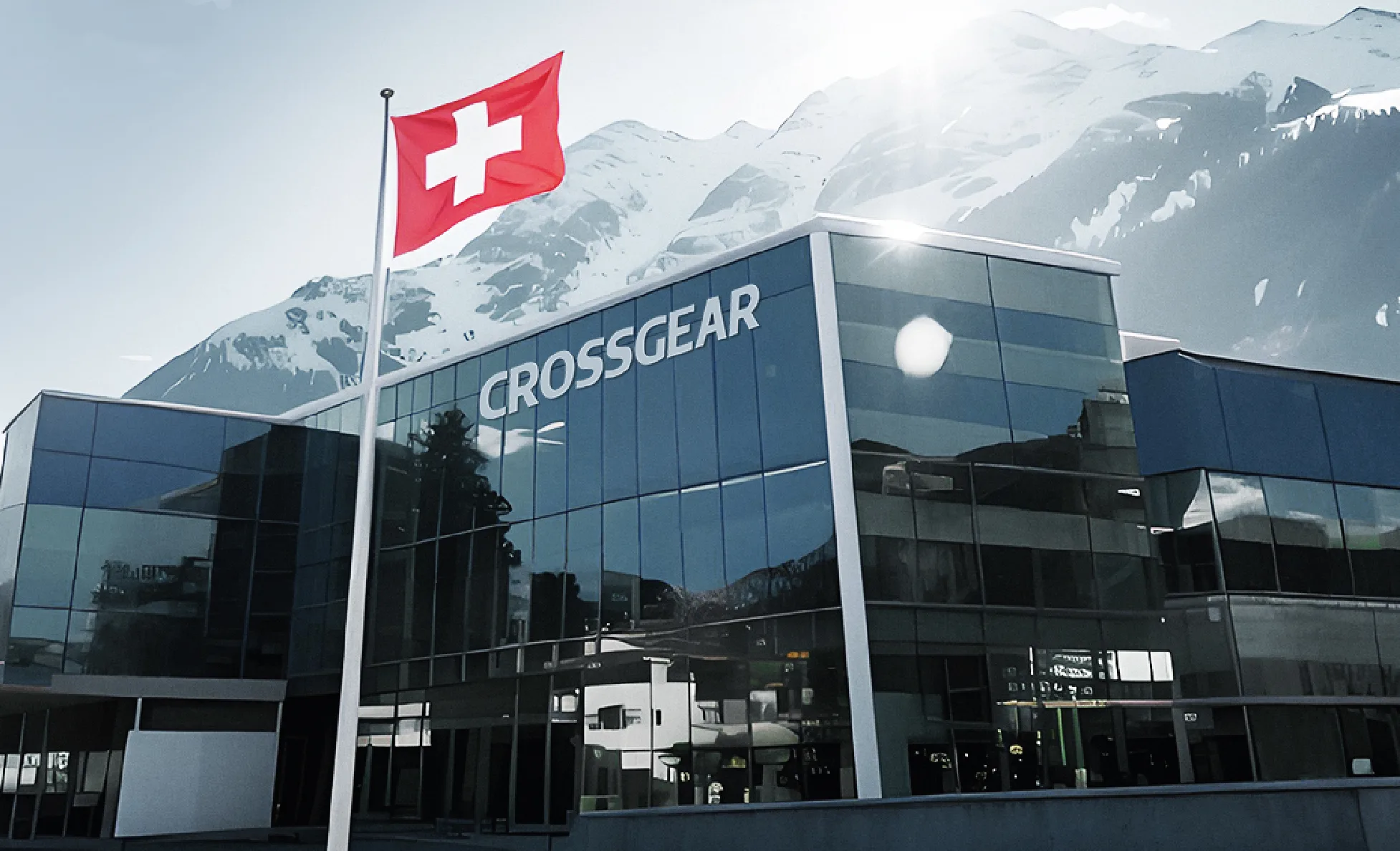 FCSI | A modern glass building labeled "CROSSGEAR" stands with a Swiss flag flying in front, set against a backdrop of snow-capped mountains under a bright sky.